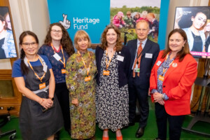 A group of people with name badges stand in front of a banner reading 'Heritage Fund'. The people include Multilingual Museum staff, volunteers, and MPs.